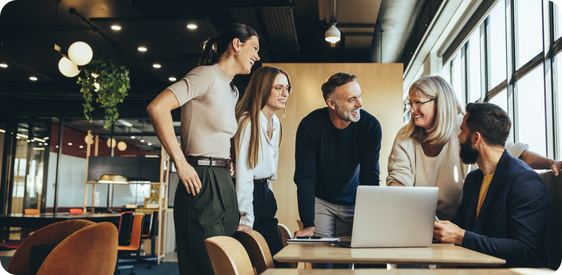 A diverse team of five professionals collaborating around a laptop in a modern office space with large windows and contemporary furniture.